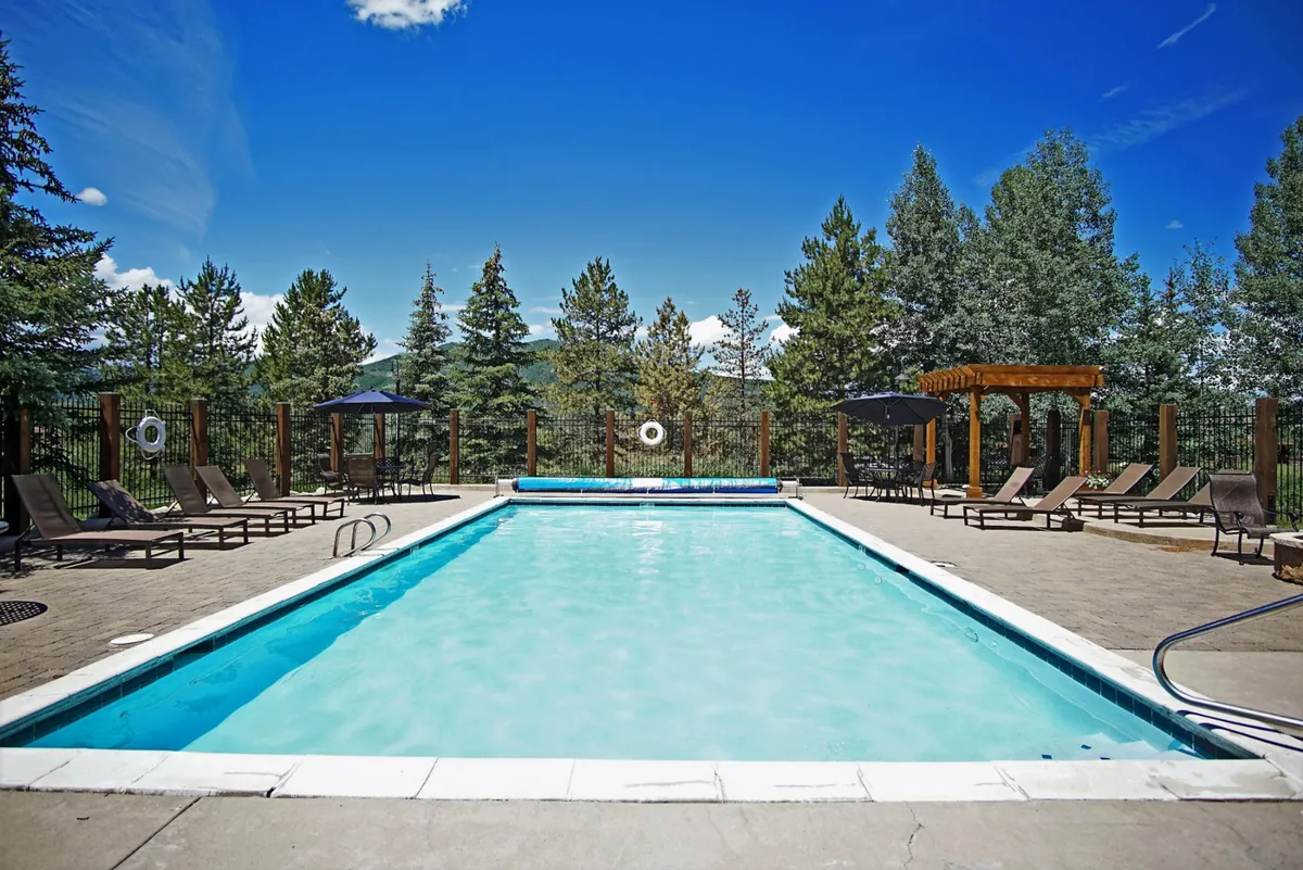 A large outdoor swimming pool surrounded by trees and lush landscaping, with lounge chairs and sun umbrellas set up poolside. The inviting water glistens under the afternoon sun, and the area is spacious enough for family gatherings, swimming, sunbathing, and social activities.