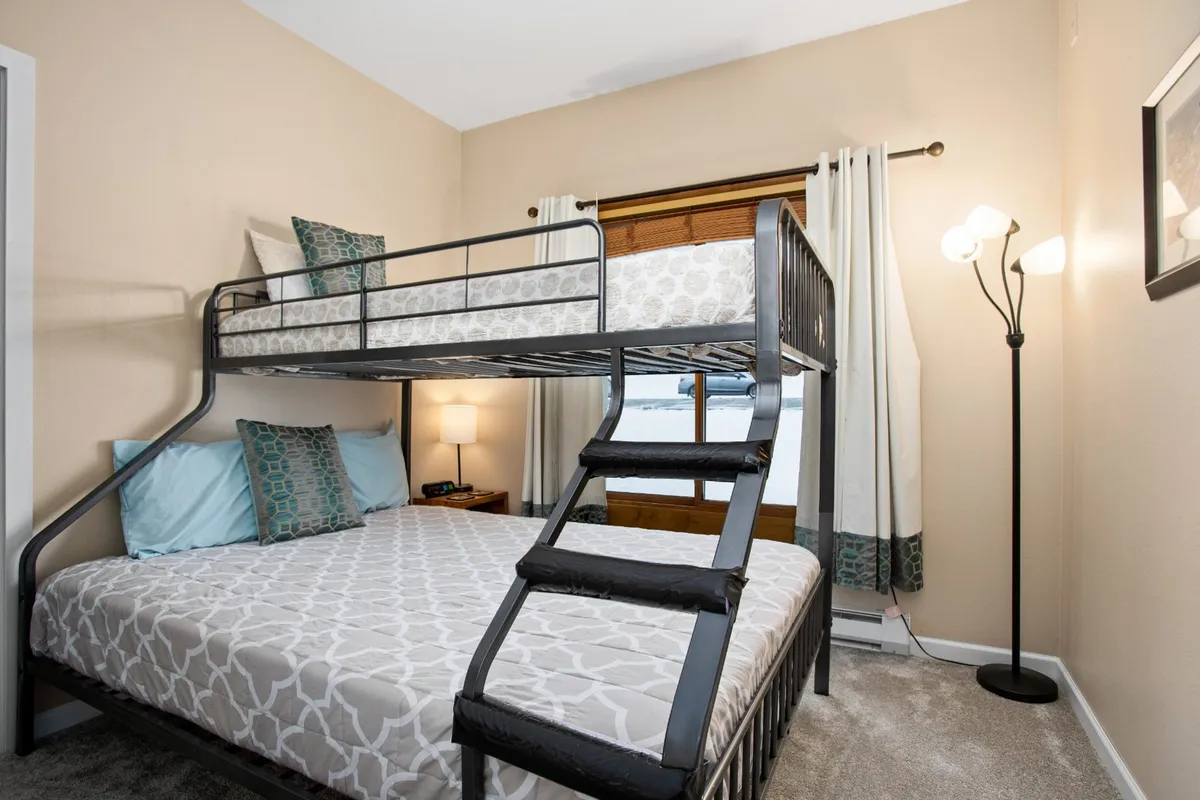 A warm, inviting bedroom scene with a bunk bed, bedside lamp, and window with curtains. The setup is ideal for kids or couples, echoing the flexibility of sleeping options described. The natural light and comforting details create a friendly, versatile space.