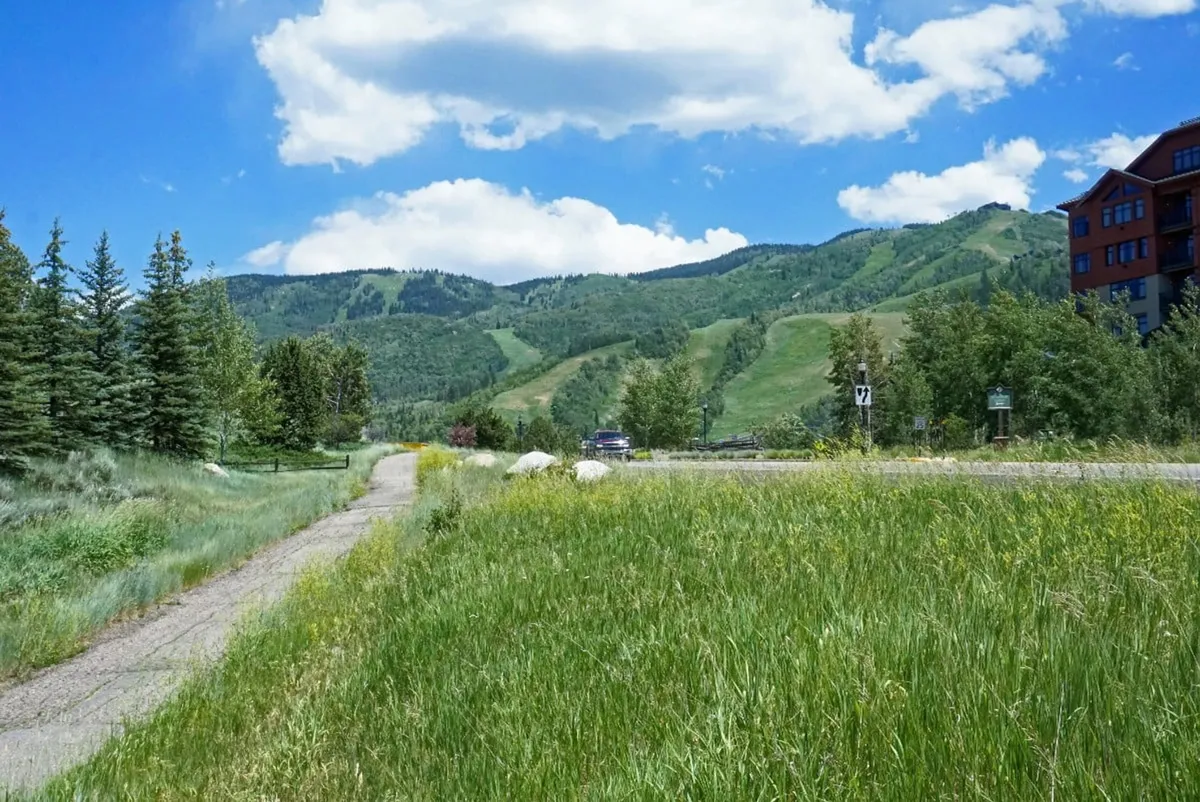 A scenic pathway leading through a grassy field toward majestic mountains, with residential resort buildings in the background. The scene suggests easy access to hiking and biking trails, and the spaciousness invites exploration and outdoor adventure.