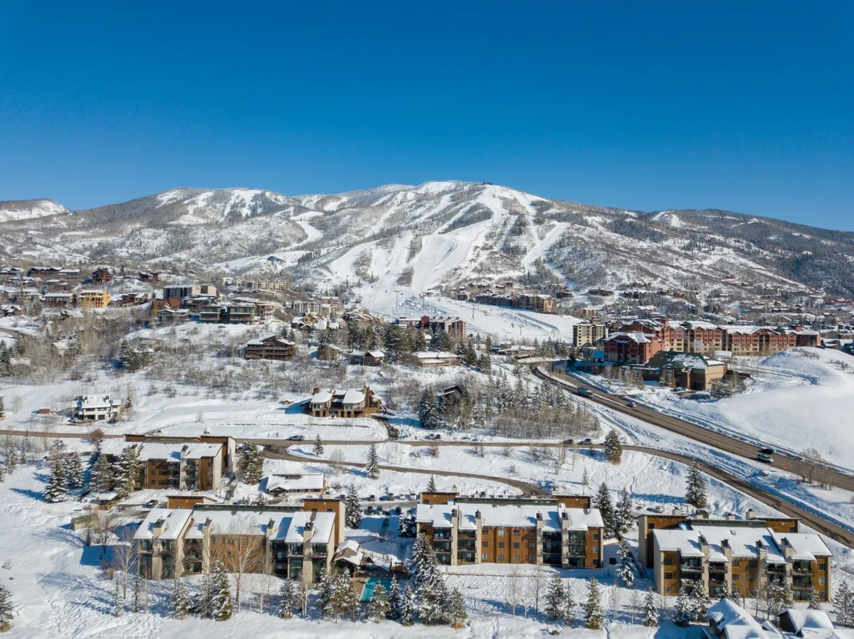 This exterior image captures snow-covered mountains, ski slopes, residential buildings, and pine trees under a crisp winter sky. It showcases the breathtaking scenery and immediate proximity to world-class skiing and snowboarding in Steamboat.