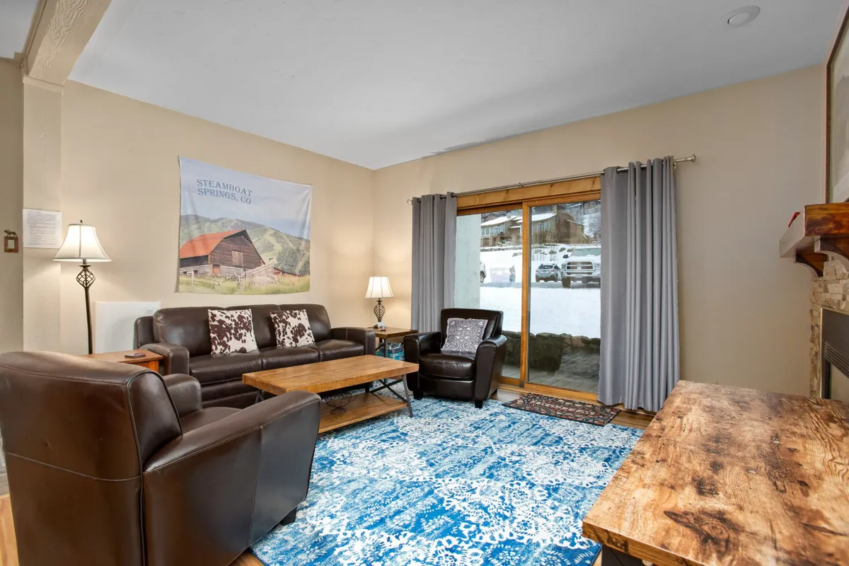 A spacious, stylish living area featuring comfortable leather sofas, a wooden coffee table, a large area rug, and a sliding glass door that leads to a private outdoor space. The layout is open and conducive to family gatherings or relaxing after a day in the mountains.