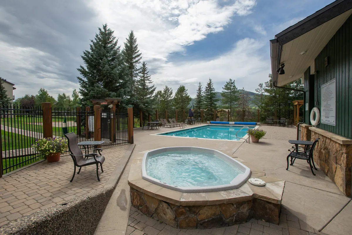 A beautiful outdoor scene showcasing a spacious swimming pool adjacent to a hot tub, surrounded by lush trees and several lounge chairs, with sunlight filtering through. The setting feels tranquil and recreational, perfect for both relaxation and social gatherings.