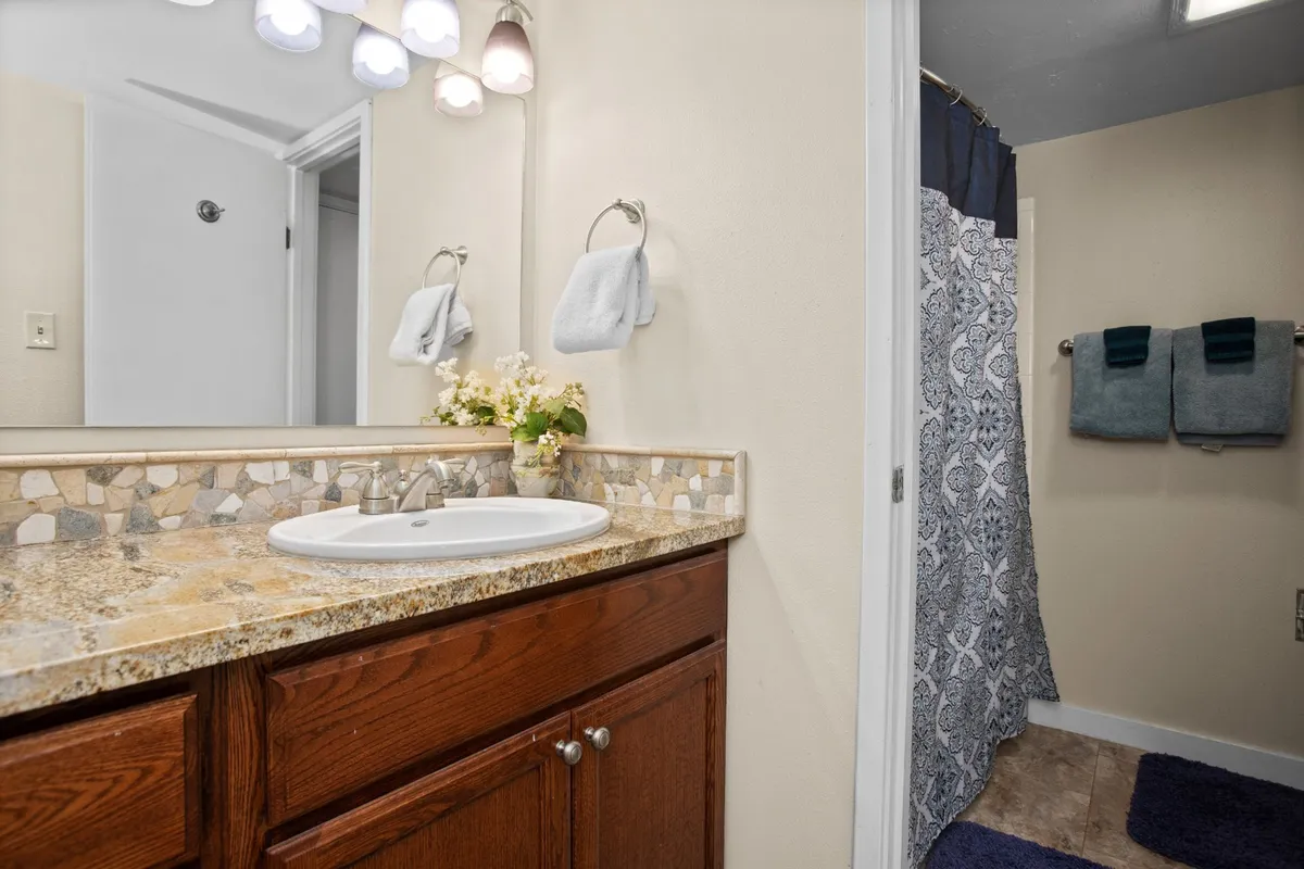 A fresh, functional bathroom space featuring a granite countertop, sink, decorative flowers, light fixtures, and a patterned shower curtain. The ambiance is both inviting and practical, making it easy to imagine morning and evening routines here.