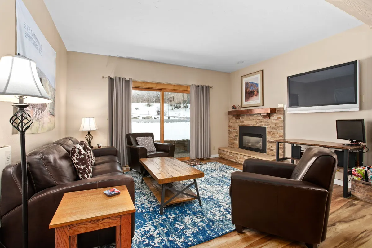 This image features a welcoming open-concept living room with plush leather sofas arranged around a cozy fireplace. A large window lets in natural light and offers a view of the private patio, while a sizable HDTV is positioned for comfortable movie watching. The overall atmosphere is warm and inviting, perfect for gatherings and relaxation.