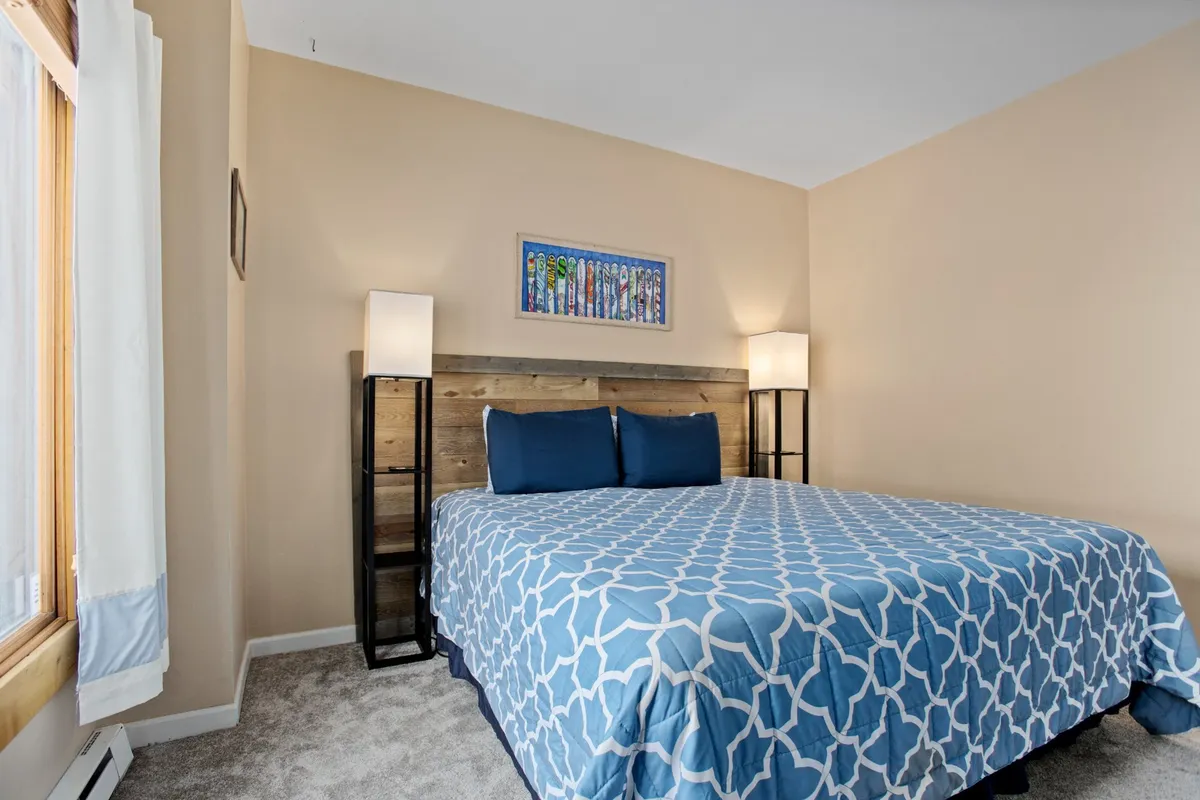 This image features a cozy bedroom with a bed adorned in blue patterned bedding, a wooden headboard, and stylish lamps on either side. Artwork decorates the wall, and the overall atmosphere is tranquil and inviting, suggesting restful nights and peaceful mornings.