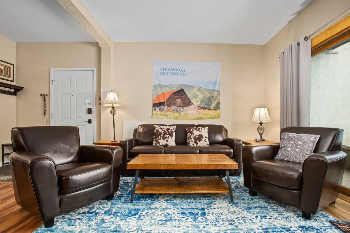 A comfortable living room setup with leather chairs, a coffee table, a wall tapestry, and soft lighting from lamps. The space radiates warmth, providing an alternative sleeping option with its sleeper sofa and a place to unwind or work.