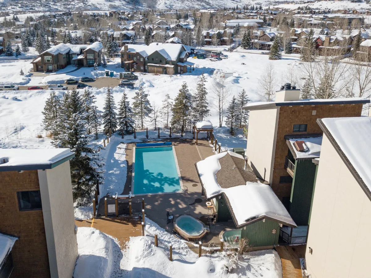 An inviting swimming pool and hot tub set within a wooden deck, surrounded by a snowy landscape and evergreen trees. The area is empty, emphasizing peace and the potential for year-round relaxation and recreation regardless of the season.