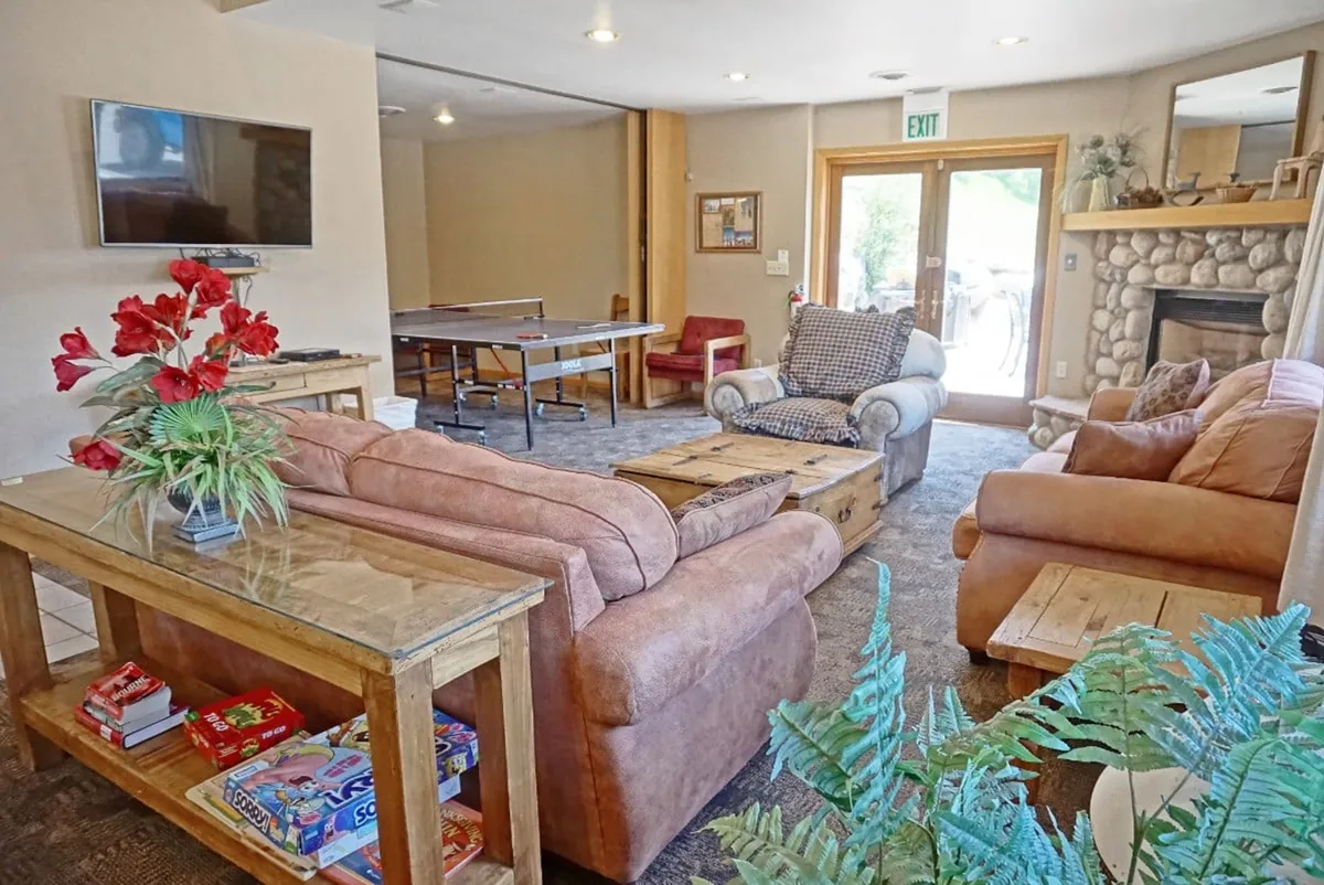 A welcoming clubhouse interior with comfortable sofas, a ping pong table, a coffee table with games, a stone fireplace, and abundant natural light. The space encourages relaxation, entertainment, and family gatherings.