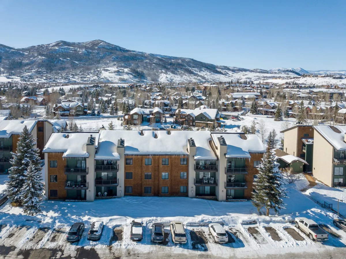A snow-covered apartment building nestled against a tranquil mountain backdrop, with the parking lot and walkways dusted in white, and distant peaks under a soft winter sky. The scene evokes the feeling of being steps away from winter adventures and cozy indoors.