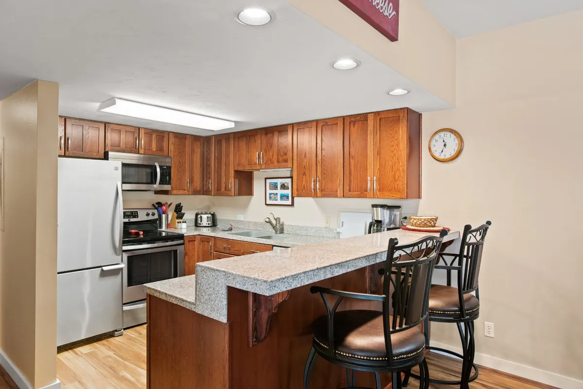 A bright kitchen featuring gleaming granite countertops, modern stainless steel appliances, warm wood cabinetry, and bar stools, offering a welcoming space for both cooking and casual dining. The kitchen is well-lit and spacious, perfect for family gatherings or entertaining guests.