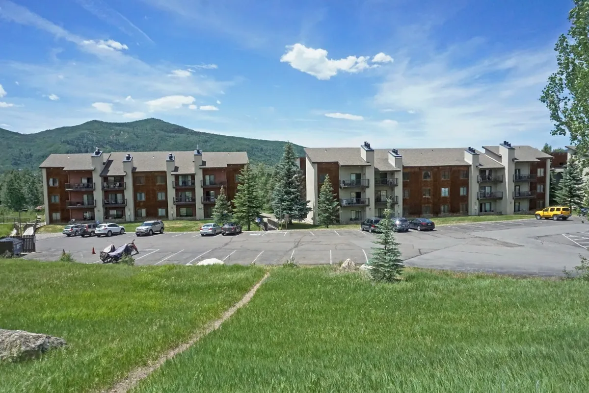 This exterior image captures the apartment buildings with a stunning mountain backdrop, green grass, and a parking lot in the foreground. The scene is open and showcases the natural surroundings as well as the ease of ground-level access.